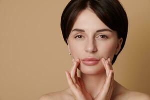 Beauty portrait of half naked European young gorgeous woman massaging her face, looking confidently at the camera isolated on beige background with copy space. Skin care, cosmetology and spa concept photo