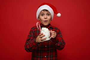 Beautiful amazed preteen boy wearing a Santa hat poses against red background with a cup of hot chocolate drink with marshmallows and striped sweet candy cane. Christmas concept with copy space for ad photo