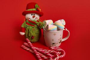 Beautiful Christmas snowman plush toy, delicious sweet sugary striped candy canes and mug with hot drink with multi colored marshmallows isolated over red background, copy space. Christmas decoration photo