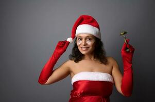 Pretty brunette in Santa carnival costume, holds jade roller for facial massage, poses smiling with beautiful toothy smile over gray background with copy space for Christmas ad of beauty spa salons photo