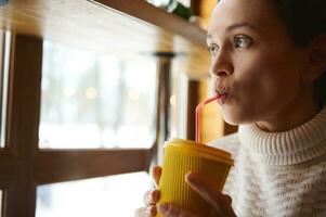 Face portrait of a young woman drinking tasty drink from a straw and looking out the window at a snow covered nature photo