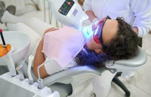 hermosa joven mujer paciente sentado en dentistas silla, con abierto boca recepción blanqueamiento tratamiento con moderno especial ultravioleta ligero lámpara para dientes blanqueo procedimiento en dental clínica foto