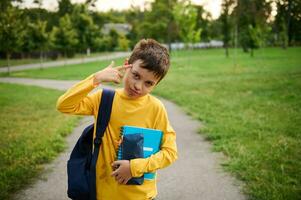 un colegial, da afuera, mirando a cámara con cansado ojos, trae su mano a su templo, imitando un pistola, como firmar de cansancio desde estudiar después un difícil colegio día en contra el antecedentes de un ciudad parque foto