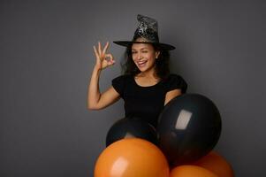 Happy joyful smiling toothy smile Hispanic woman wearing wizard hat, dressed in witch carnival costume for Halloween party, shows OK sign, poses against gray background with black balloons, copy space photo