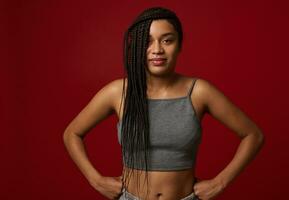 Waist length of a pretty woman of African ethnicity, with stylish dreadlocks, putting hands on waist and cute smiles confidently looking at camera, isolated over red background photo