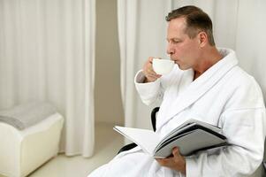 Handsome mature 50 years old European elegant man dressed in white terry bathrobe, drinking healthy herbal tea and reading magazine sitting on armchair in private room of luxury wellness spa centre photo