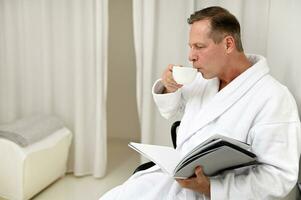 hermoso maduro 50 años antiguo europeo elegante hombre vestido en blanco terry bata de baño, Bebiendo sano herbario té y leyendo revista sentado en Sillón en privado habitación de lujo bienestar spa centrar foto