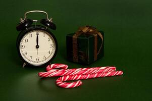 Close-up of black alarm clock with midnight on the dial, sweet striped lollipops, candy canes and Christmas gift in glitter wrapping paper and golden bow isolated on green background with space for ad photo