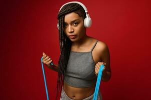 Determined African sportswoman in gray sportswear exercising with an elastic fitness strap during routine workout, isolated over red background with copy space. Body positivity, fitness, sport concept photo