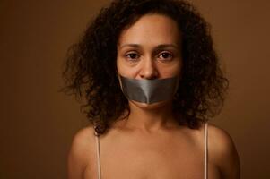 Close up portrait of frightened desperate mixed race woman with sealed mouth looking at camera with hopelessness, isolated on dark beige background. International Day to End Violence against Women. photo