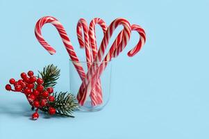 Beautiful minimalistic simple composition with Christmas lollipops in transparent glass and snowy branch of pine with red berries, holly, on blue background with copy space for advertising photo