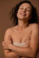 Delighted confident dark-haired curly woman with closed eyes and skin flaws in beige underwear smiles toothy smile posing against beige background with copy space for ads photo