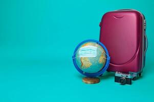 Suitcase, vintage camera and globe - planet Earth concept - with protective medical mask isolated on blue background with copy space for ad. Tourism safety and travel concept during pandemic photo