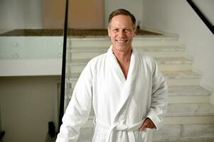 Portrait of a confident handsome middle aged man wearing terry bathrobe and smiling toothy smile looking at camera standing on the stairs of health spa while enjoying relaxing male beauty procedures photo