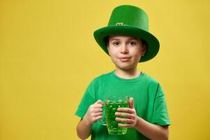 Little boy in a green leprechaun Irish hat holds a glass with a green drink. Saint Patrick day. Isolated on yellow background.. photo