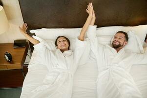 Happy young married couple, newlyweds, gorgeous wife and husband, lovers stretching in the bedroom after waking up, touching each other's hands and smiling, enjoying the privacy on their honeymoon photo