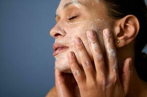 Head shot of beautiful woman with perfectly healthy skin, holding hands on her face, performing smoothing massage, removing makeup and cleansing face with exfoliating scrub and facial foam cleanser photo