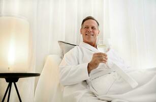Handsome middle aged Caucasian man in white terry bathrobe holds a drinking glass with water, smiles looking at camera, enjoying his day off at private massage room of a luxurious wellness spa centre photo