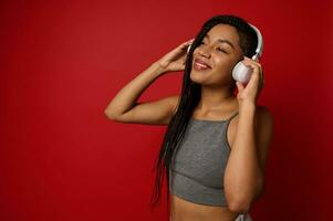 Beautiful awesome happy young African woman putting wireless headphones on her head and enjoying listening to music on red background with copy space photo