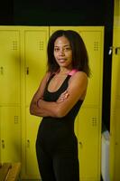 Confident smiling African sportswoman with perfect slim body in black sportswear stands with crossed arms in front of yellow lockers in gym dressing room, looking at camera photo