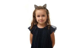 Portrait of cute little witch smiling girl wearing a hoop with cat ears, dressed in stylish carnival dress, looking at camera posing with crossed arms against white background, copy space, Halloween photo