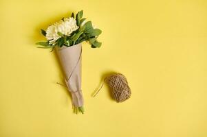 Flat lay floral arrangement of a delicate beautiful bouquet of autumn asters flowers in kraft wrapping paper tied with a rope and a skein of rope on a yellow background with space for text photo