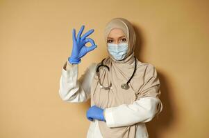 Beautiful Muslim doctor in medical mask covered head in hijab posing on beige background with copy space and gesturing OK with her fingers photo