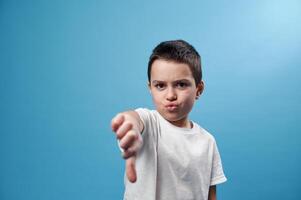 Schoolboy showing a thumb down at camera. Facial emotions on blue background with copy space photo