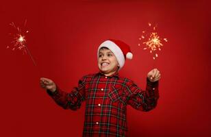 Christmas and New Year celebration concept. Funny handsome child boy in Santa hat with sparklers against red colored background with copy space for ad photo