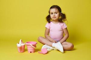 Cute little girl wearing summer clothes , sitting near set of beach toys, looking at camera while posing over yellow background photo