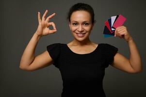 Beautiful Hispanic woman smiles with toothy smile and poses against gray background with many discount or credit cards in her hands showing ok sign looking at camera. Shopping concept at Black Friday photo