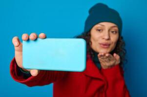 Focus on the hand of a blurred pretty woman in bright red warm cozy woolen coat and green hat sending air kisses while making selfie and talking video call against blue background with copy ad space photo