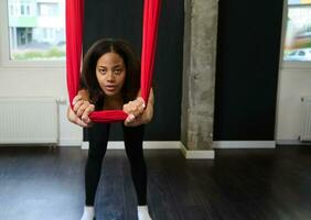 Slim body beautiful African sporty woman in black activewear stretching her arms and back muscles using red hammock during aerial flying yoga photo