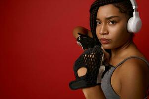 Concentrated young African athletic woman boxer in headphones and red boxing gloves, looking at camera making a direct hit, isolated over color background with copy space. Contact martial art concept photo