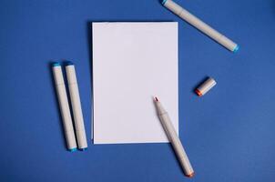 Two-color flat lay composition with watercolor markers or felt-tip pens with white blank sheet of paper with copy space isolated on blue background. photo