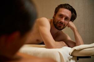 Handsome middle aged European man lying on a massage table, smiles looking at camera, getting ready to receive professional body massage therapy at wellness center. Newlyweds couple at health spa photo