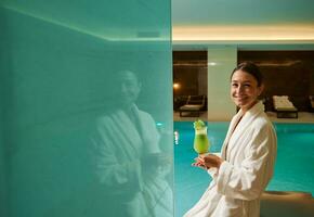 Middle aged pretty woman in white bathrobe stands by the pool in a wellness spa centre, holds freshly squeezed vitamin juice in hands, smiles looking at camera. Healthy lifestyle, recreation concept photo