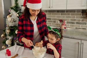 Beautiful Hispanic woman, happy loving mom and her beloved cute daughter in red checkered outfit having fun together, preparing dough for cooking Christmas bakeries at home kitchen. photo