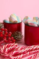 High angle view of cropped image of Christmas items, striped sugary candy canes, holly and cup with hot drink and wet colorful marshmallows on pink colored background with copy space for Christmas ad photo