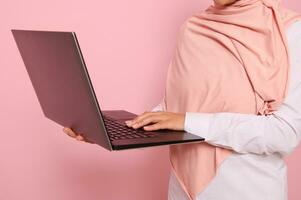 Close-up portrait of a woman wearing pink hijab standing against colored background with an open laptop in hands and typing text.Concept of successful Muslim programmer woman, worker at IT technology photo