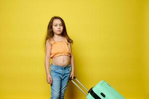 Adorable Caucasian kid girl in trendy summer clothes, with light green stylish suitcase, isolated on yellow background photo