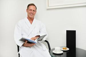 Handsome charming mature Caucasian man in white terry bathrobe sitting on armchair with a magazine in his hands, smiles toothy smile looking at camera, relaxing at luxurious wellness spa centre photo