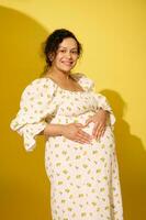 Beautiful pregnant woman in sundress holds her hands on her big belly, making a shape of heart, smiles looking at camera photo