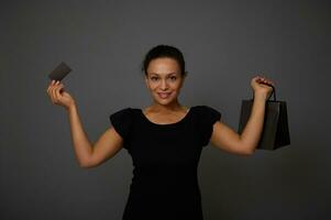 Isolated portrait on gray background with copy space of an attractive smiling woman dressed in black and holding a discount credit card and a shopping bag, looking at camera. Black Friday concept photo