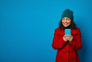 Isolated portrait on blue background of a cheerful awesome happy pretty woman wearing bright red coat and warm green woolen hat holding telephone and rejoices looking aside .Copy ad space photo