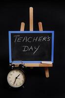 Close-up of a chalk board with lettering Teacher's Day, standing on a wooden table easel, next to black alarm clock, isolated on black background with copy space. photo