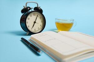 An open notebook with schedule, ink pen, black alarm clock lie on blue surface. Color background with copy space. Time management, deadline and schedule concept photo