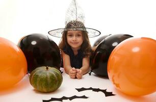 tradicional evento, Víspera de Todos los Santos fiesta concepto. hermosa pequeño bruja niña en un magia sombrero acostado en un blanco antecedentes con Copiar espacio siguiente a negro hecho a mano sintió murciélagos, calabaza y naranja negro globos foto