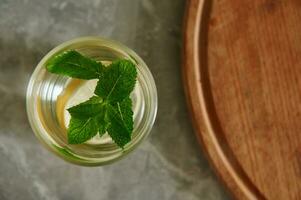 Healthy mint lemon drink with mint leaves and sliced lemon in a glass next to a cut round wooden board on a gray kitchen surface. Flat lay photo