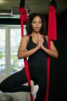 Attractive young African woman practicing flying yoga and sitting in lotus position, hanging in a hammock and making a prayer gesture. Aerial aero fly fitness. photo